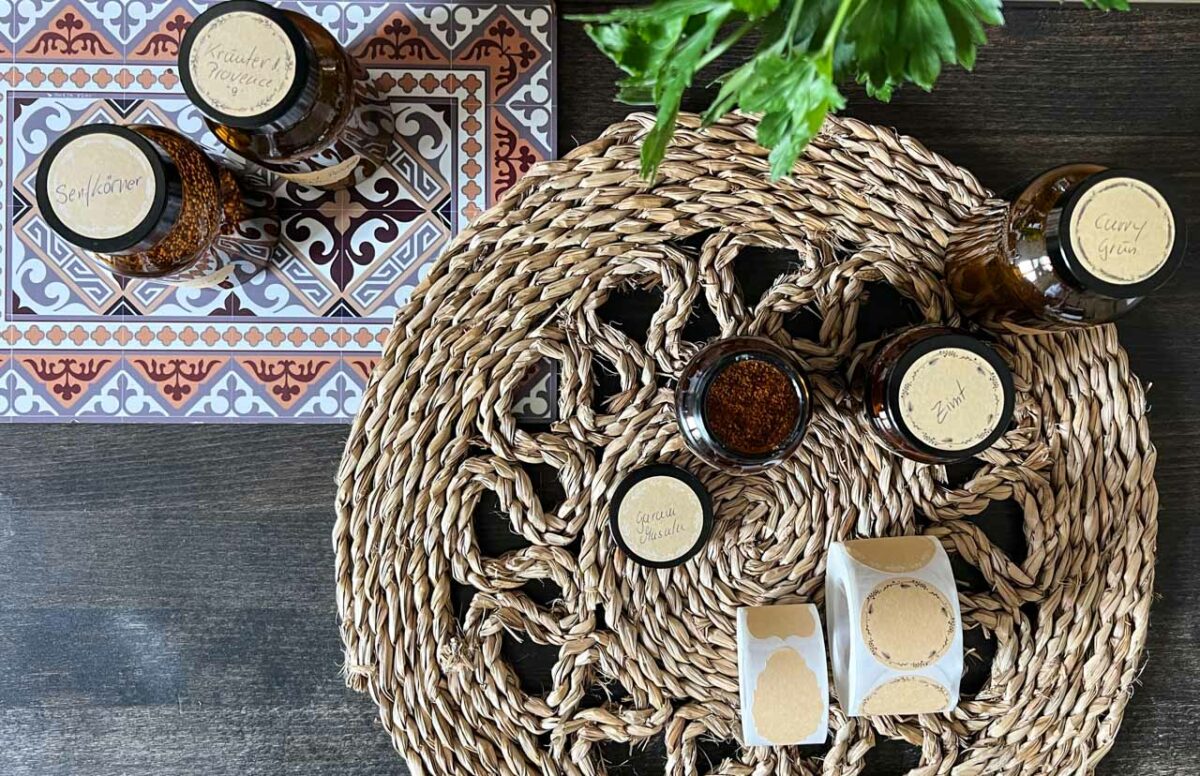 Apothekengläser von oben auf einem geflochtenen Tischset, mit runden Kraftpapier-Aufklebern beschriftet, neben einer Rolle Aufkleber und dekorativen Fliesen.