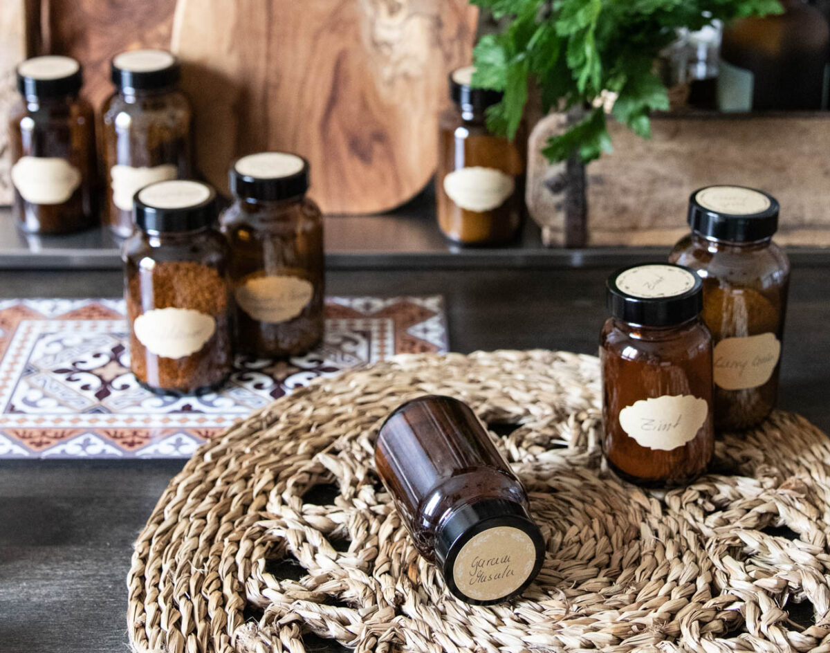 Braune Apothekengläser mit Gewürzen auf einem geflochtenen Platzset und dekorativen Fliesen im Hintergrund.